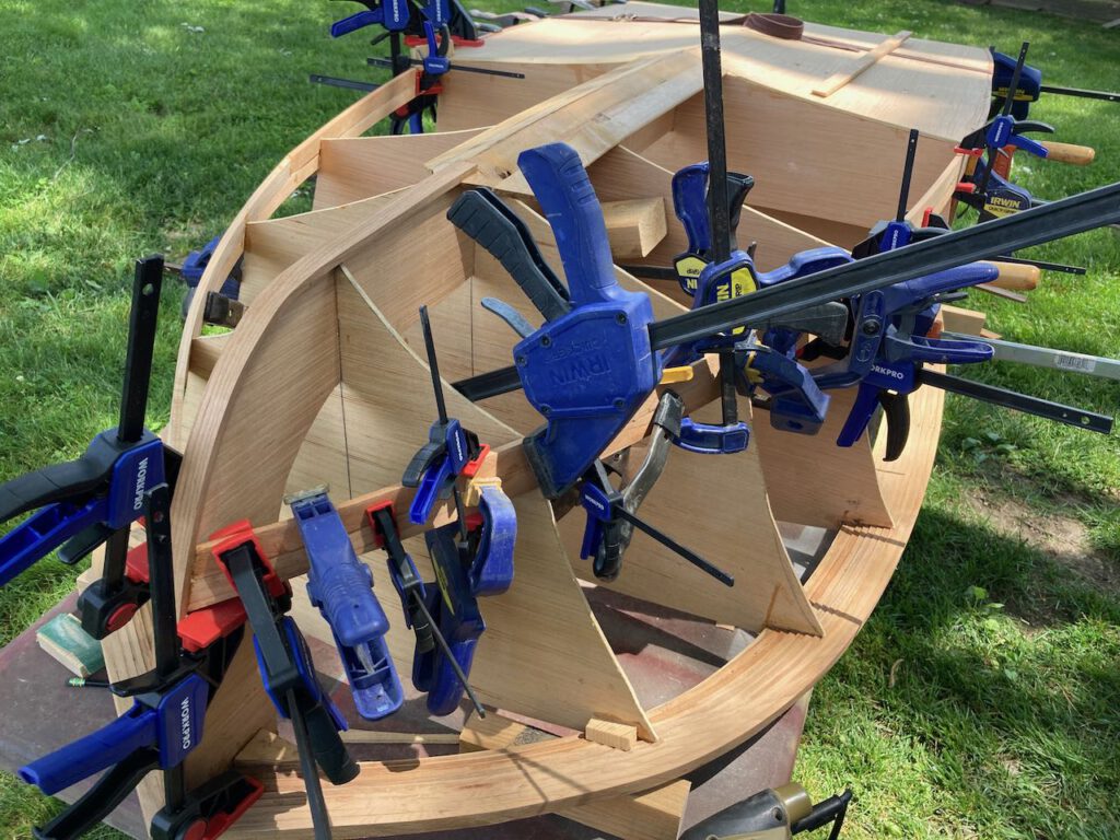 Multiple clamps secure the first couple strips of wood that make up the sides of the hull of this wooden boat under construction, seen upside down on a table outdoors.