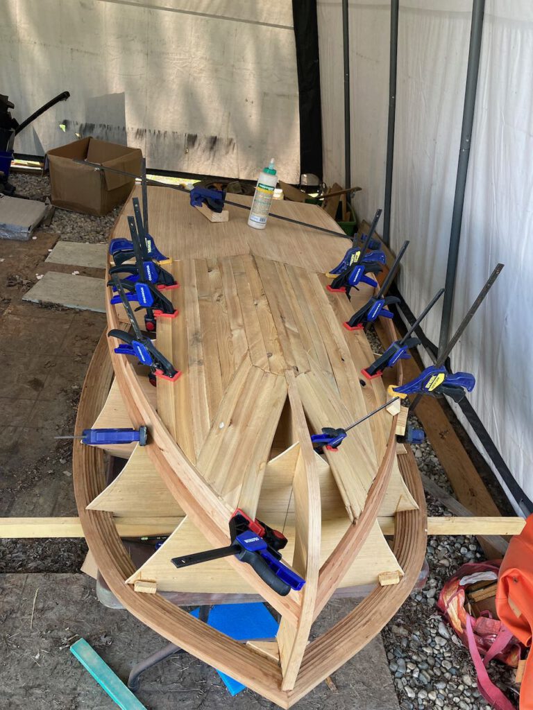 Clamps on the hull of a wooden boat under construction. The boat is upside down with a series of clamps holding strips of wood forming the bottom of the vessel.