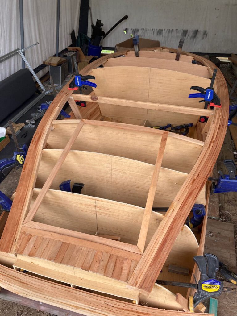 Wooden boat under construction. Looking down from above at the aft deck of the vessel. Framing and some decking is in place outlining the shape of the boat.