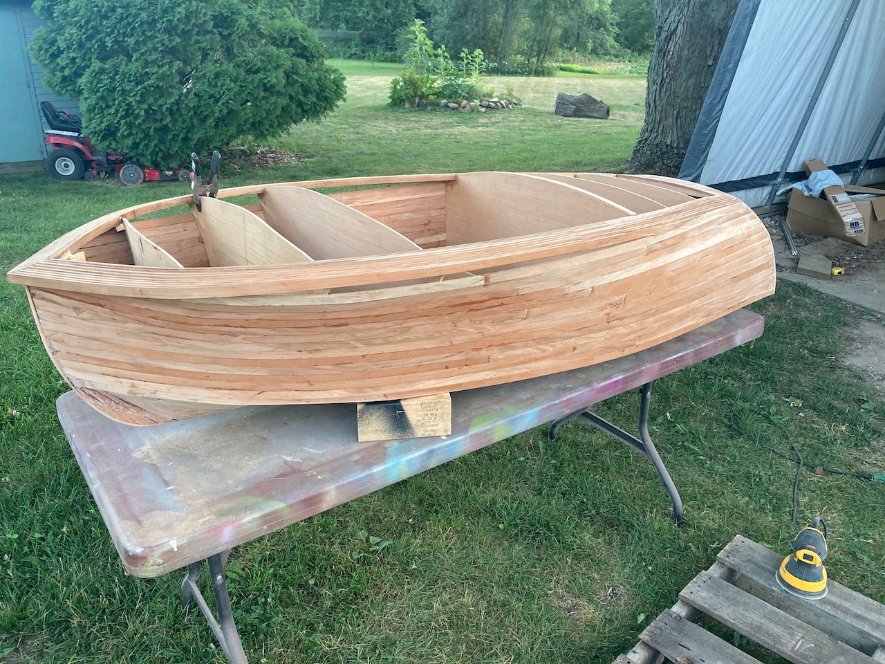 This small boat under construction is resting on a table outdoors. The hull is not quite complete, but parts of it have been roughly sanded.
