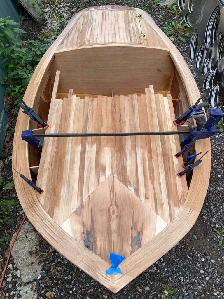 Wooden boat under construction, seen from above. Clamps hold in place strips of cherry wood making up the foredeck.