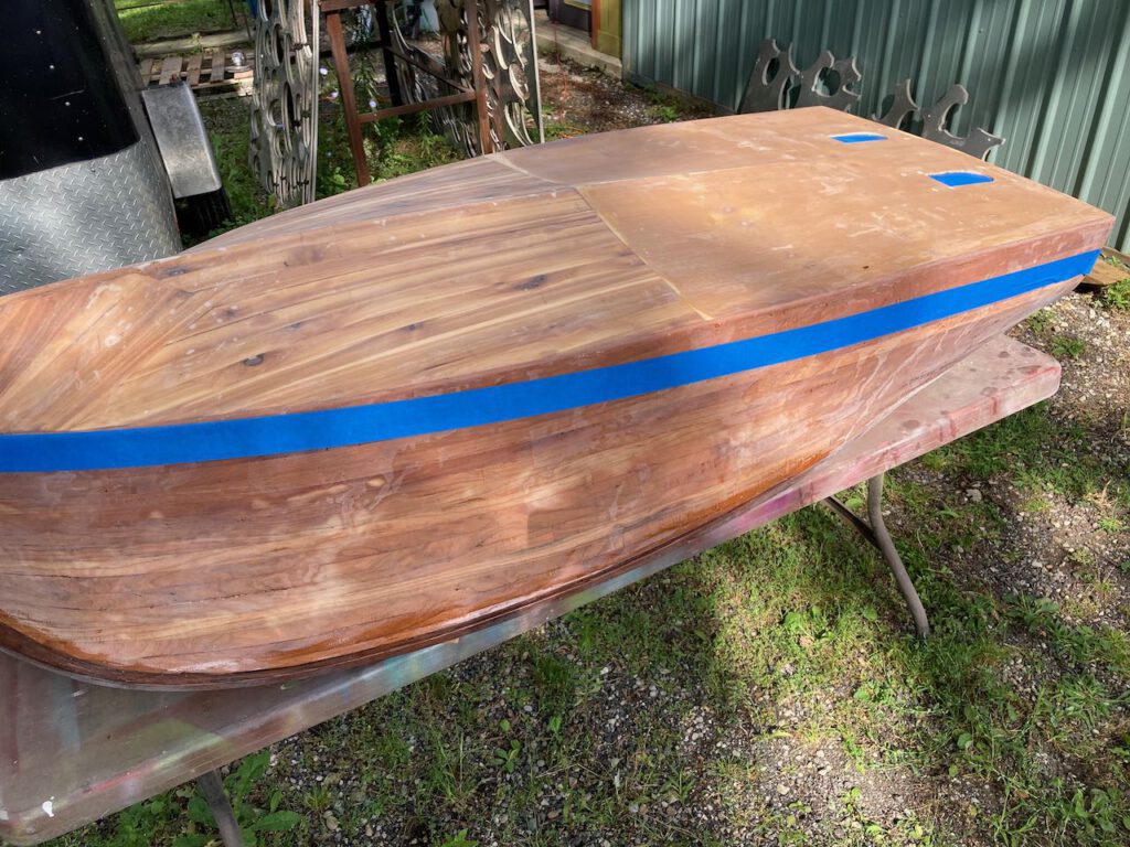 Wooden boat under construction. The hull is upside down and a strip of blue painters tape marks the water line.
