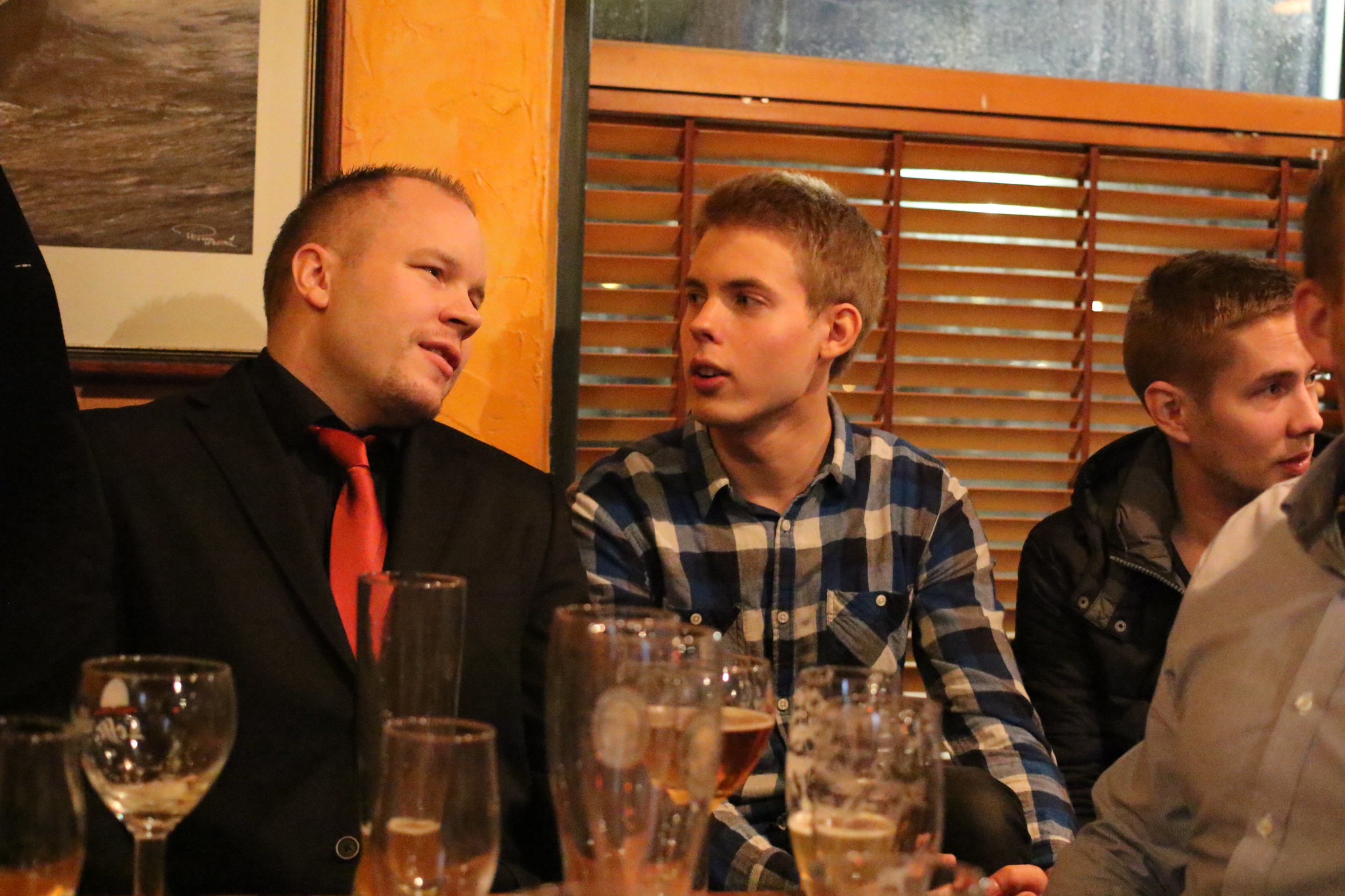 a group of men sitting and talking around a table full of partially empty glasses. The atmosphere appears to be a crowded restaurant. The men are talking with each other but appear partially distracted by something out of view.